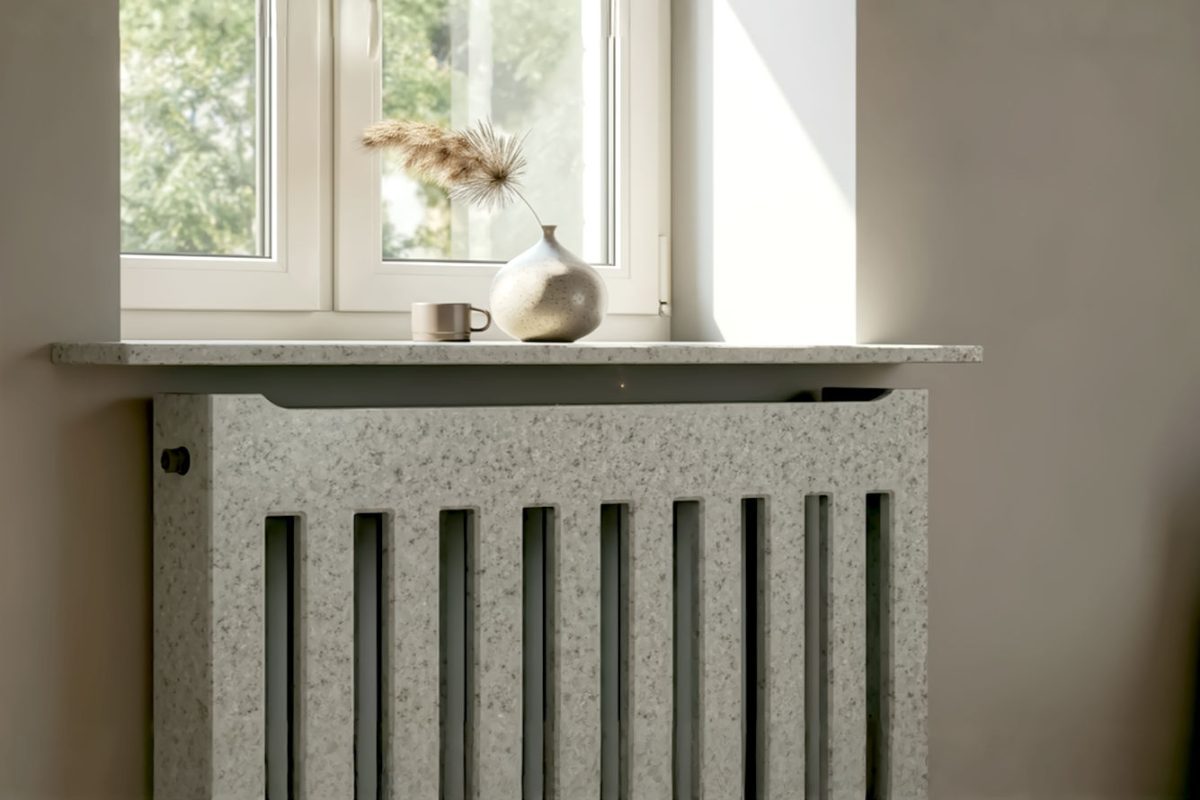 A professionally lit, close-up shot of a modern living room featuring a beautifully polished light grey granite radiator cover under a window, with a stylish vase and a small plant on top. The granite's subtle sparkle should be visible, contrasting with a soft, neutral wall.